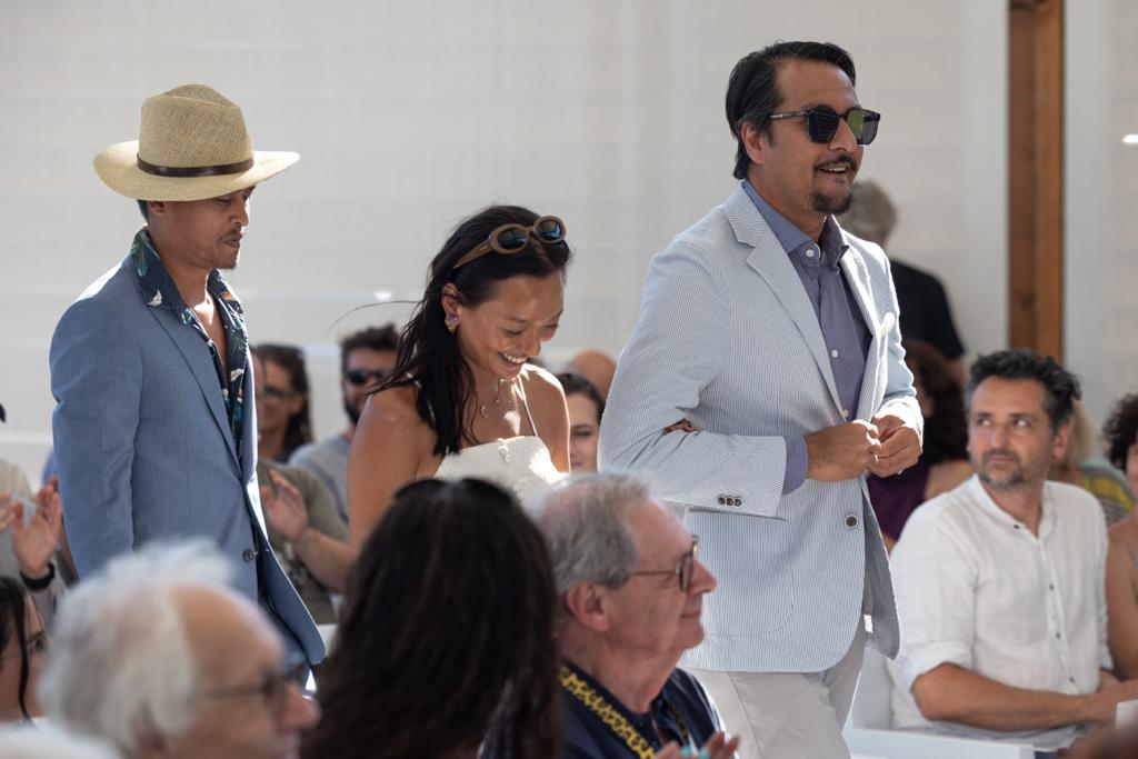 Three people are standing and walking in a sunny, public event, with spectators seated around them.