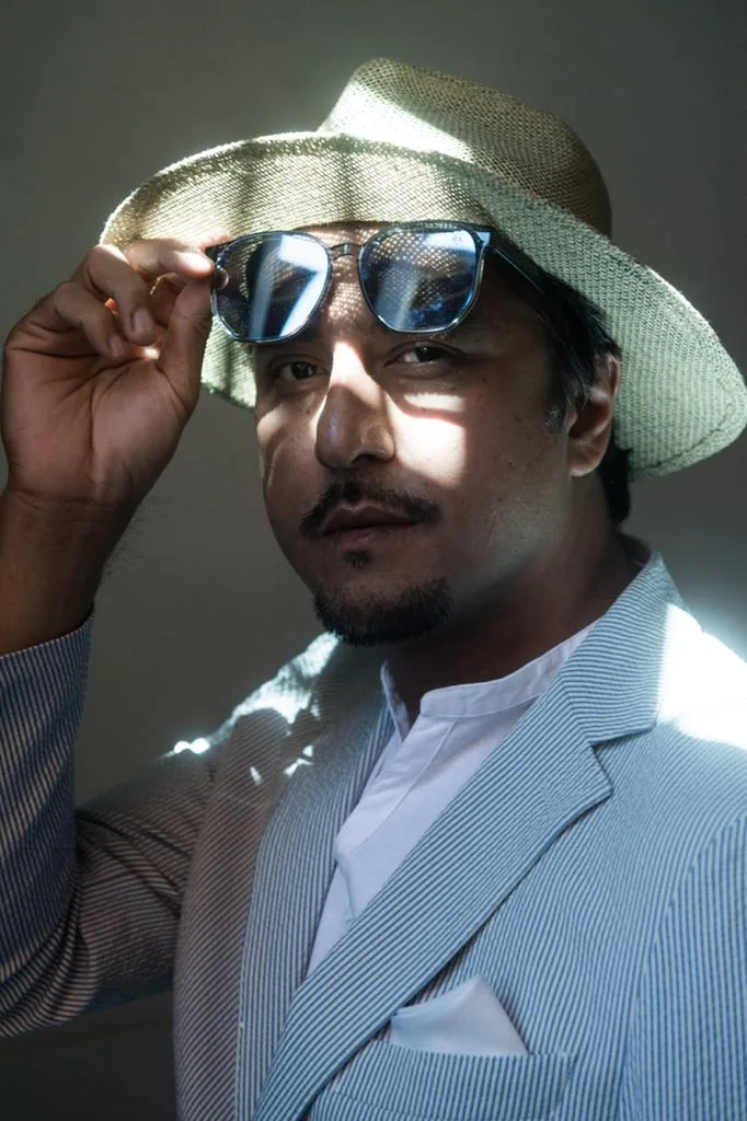 A man in a pinstripe suit and sunglasses tipping his straw hat.