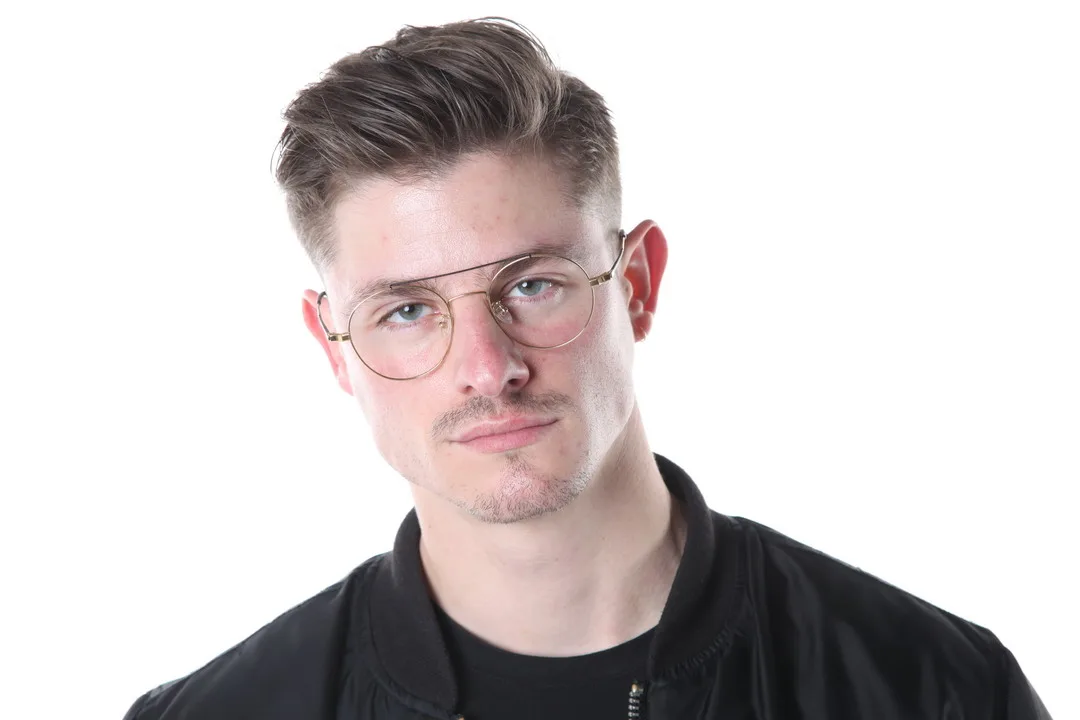 A man with glasses and a black jacket is posing with a slight head tilt against a white background.