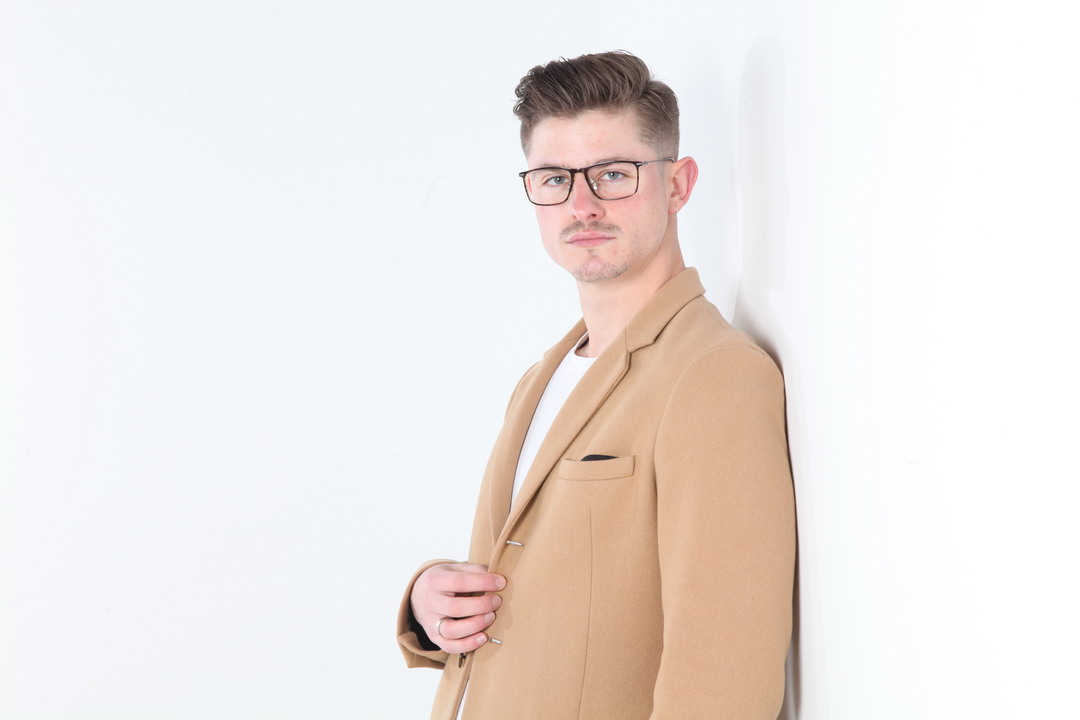 A man in glasses wearing a tan blazer poses against a white background.