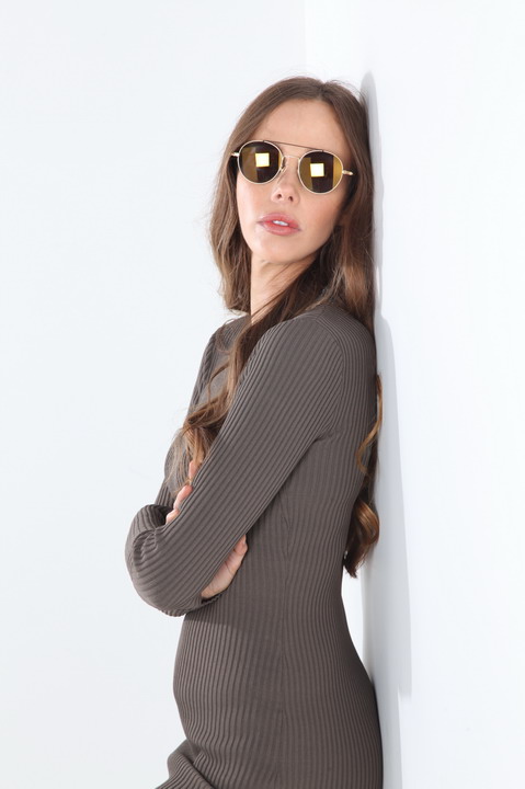 A person wearing sunglasses and a long-sleeve dress is standing with crossed arms against a white background.